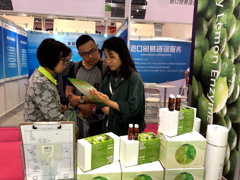 來賓在展位看綠色 DM，桌上有檸檬酵素禮盒與試飲小瓶