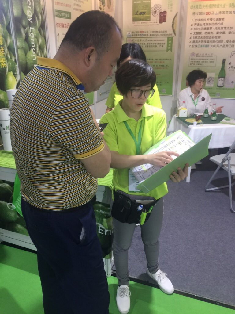 2019 上海國際酵素產業博覽會：白襯衫女士桌前解說，藍襯衫訪客手持試飲杯品飲