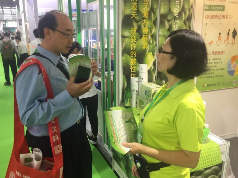 2019 上海國際酵素產業博覽會：螢綠制服成員向掛紅色飲料授帶的訪客遞型錄解說，背景綠檸檬巨幅看板