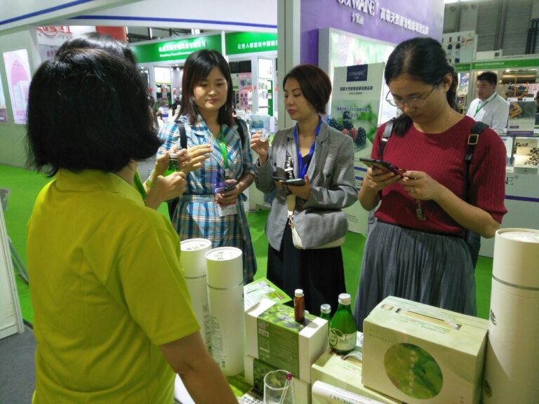 2019 上海國際酵素產業博覽會：白襯衫女性人員坐桌前，將綠玻瓶酵素原液倒入小杯為訪客品飲，兩位女性訪客分坐兩側观看；背景為清大技術指導、L99 產品與投保 3000 萬產品責任險海報